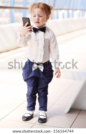 Red hair little cute girl with bow-tie stands and looks at cell phone.
