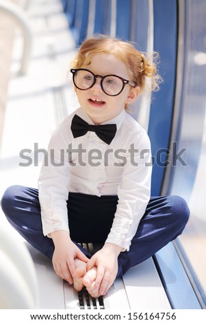 Little cute barefoot girl in glasses sits on floor in gallery near window.