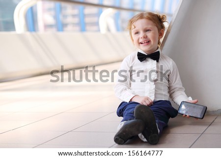 Red hair little cute girl with cell phone sits on floor and laughs.