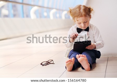 Little cute barefoot girl in white shirt sits on floor and playing with tablet pc in gallery.