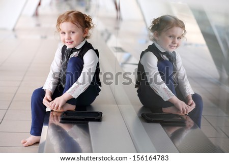 Little barefoot girl in tie sits on floor with tablet PC in gallery near glass wall.