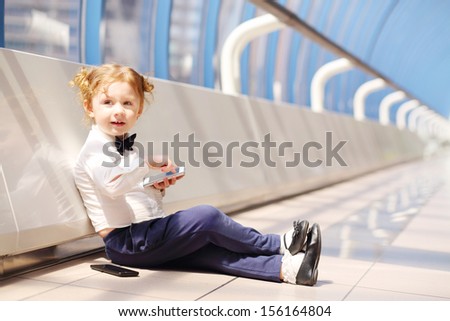 Little Cute Girl Sits On Floor With White And Black Cell Phones In Long Gallery.