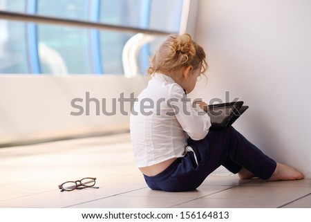 Back of little cute barefoot girl sitting on floor and playing with tablet pc in gallery.