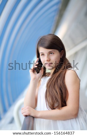 Girl in white blouse stands in long light gallery and talks by mobile phone.