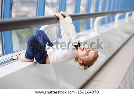 Little cute red hair girl in bow-tie hangs on handrail in gallery near window.