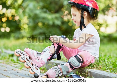 Little girl in protective equipment and rollers sits on curb of walkway in park, fastening knee-pad