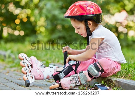 Little girl in protective equipment and rollers fastens knee-pad, sitting on curb of walkway in park.