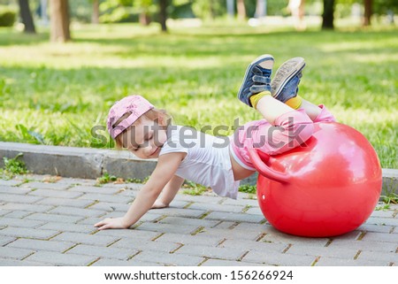 Little girl plays with red ball for jumping on  walkway in summer park, hands on ground, legs on ball