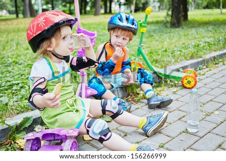Children scooterists rest sitting on curb of walkway in park, focus on girl