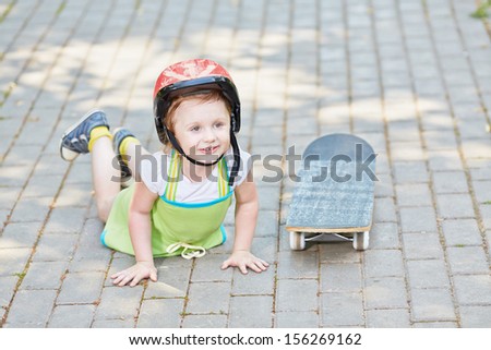Little girl in protective helmet lies on park path near to skateboard, leaning hands on ground.