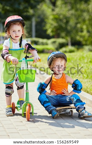 Little skateboarder sits on skateboard with his arms akimbo ,little girl with three-wheeled scooter stands next to him