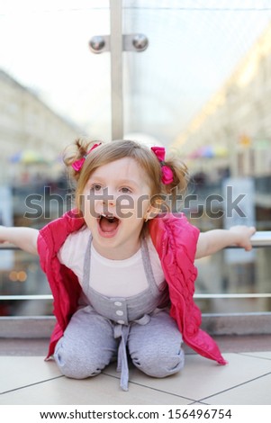 Little cute girl sits on floor, holds on to railings and shouts in mall.