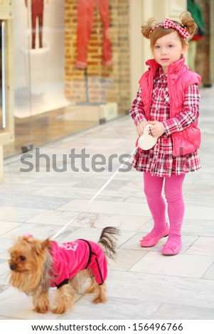 Little beautiful girl in dress leads small dog on leash in clothes in store.