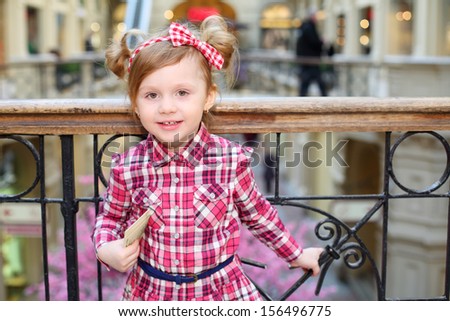 Little cute girl with papers stands near wrought railings and smiles.