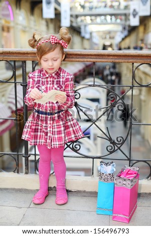 Little cute girl with papers stands near wrought railings and bags.