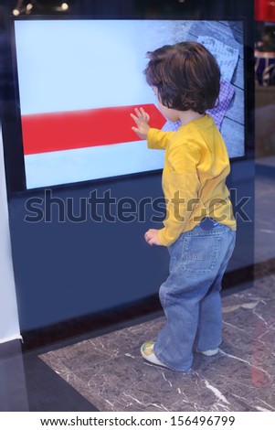 Back of little boy in jeans touching big advertising display at mall.