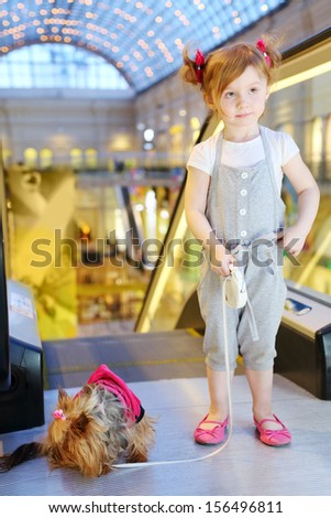 Little pretty girl with dog on leash stands next to escalator in mall.