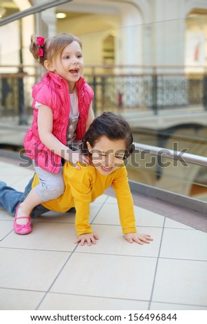 Little cute girl and boy play on floor in big mall. Girl saddled boy.