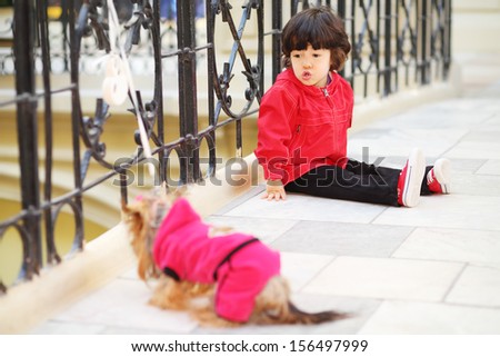Little cute boy sits on floor near wrought railings and talks with linked small dog in store. Focus on boy.