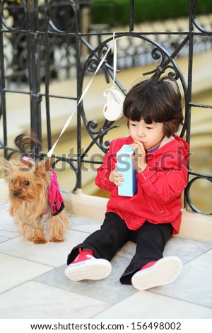 Little cute boy sits on floor and drinks juice near wrought railings and linked small dog in store.