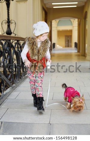 Little beautiful girl held on leash dog in pink clothes in large store.