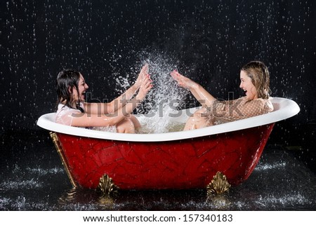 Two wet girls have fun and splashing water in the bathtub under the spray