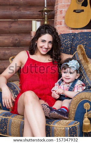 Mother with little daughter sitting on armchair in a log house with a guitar on the wall