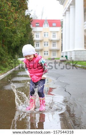 Happy little girl in a pink vest and rubber boots jumps into a puddle