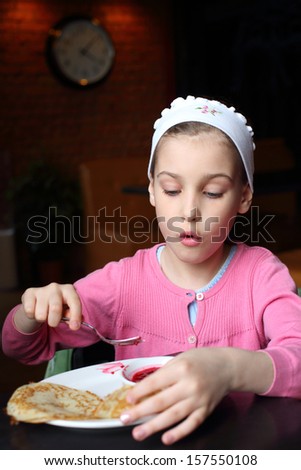 Girl in a pink jacket and a headband eating pancakes with jam in a cafe
