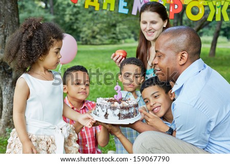 Family of six looks at birthday cake with burning candle on it, happy birthday sign behind theirs backs