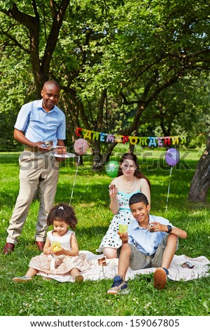 Family of four eats birthday cake sitting on rag matting on grass in park, happy birthday sign behind theirs backs