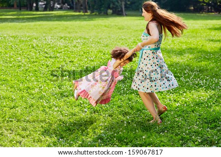 Mother turns little daughter in circle holding her hands on sunny grassy lawn in park