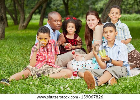Family of six sits on rag matting and eats fruits and berries in summer park