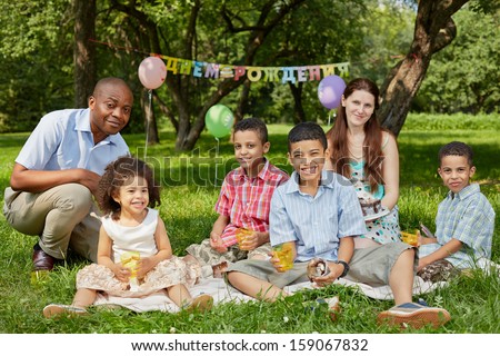 Family of six eats birthday cake sitting on rag matting on grass in park, happy birthday sign behind theirs backs