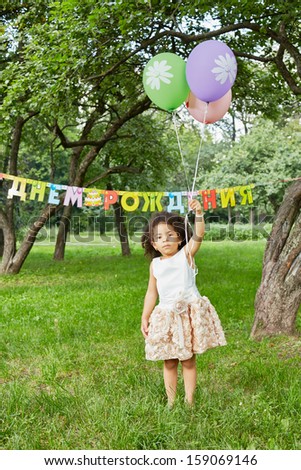 Little girl stands in park, holding three air balloons in her raised hand, happy birthday sign behind her back