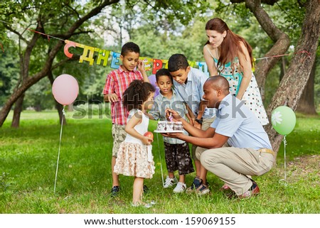 Father lights candle on birthday cake with lighter, family of five looks at it, happy birthday sign behind their backs