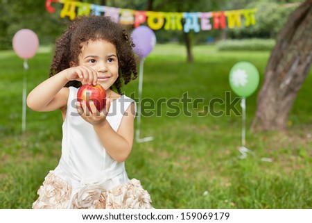 Little girl stands in park, holding big red apple in her hands, happy birthday sign behind her back