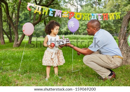 Little girl stands in park, holding big red apple in her hand, father gives her birthday cake, happy birthday sign behind her back
