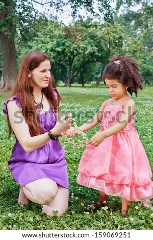 Mother stands on one knee on grass in park and gives flower wreath to her little daughter