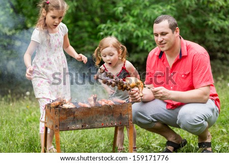 A Family Of Three Making Barbecue On The Grill On Nature, Little Girl Blowing On A Skewer With Mushrooms.