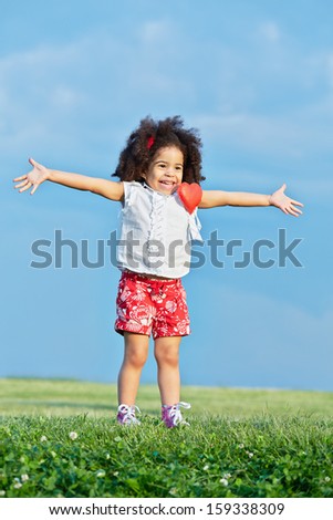 Little girl in red shorts and white t-shirt with pinned red heart stands on grassy meadow with her arms outstretched to sides