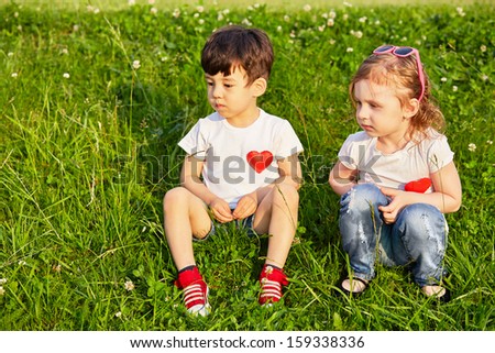 Two little children sit on grassy slope, red hearts pinned to theirs shirts