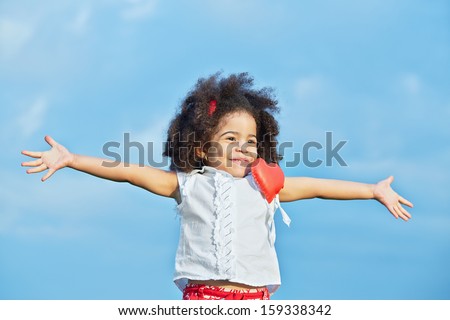 Half-length portrait of smiling little girl in red shorts and white t-shirt with pinned red heart with her arms outstretched to sides