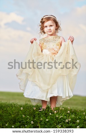 Little girl in beautiful beige gown and crown on head stands  on grassy meadow, lifting up slightly hem of external skirt