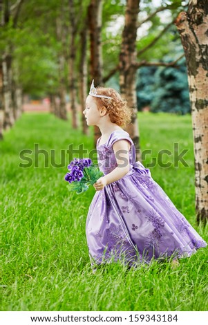 Little girl in beautiful gown and crown on head with violet flower in her hand runs on grassy lawn in park