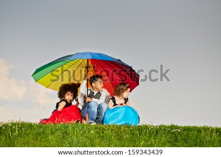 Little boy and two little girls hide from rain sitting squatted under big colorful umbrella