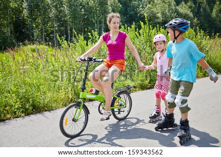 Mother rides bicycle and her children ride rollers on sunny day in park