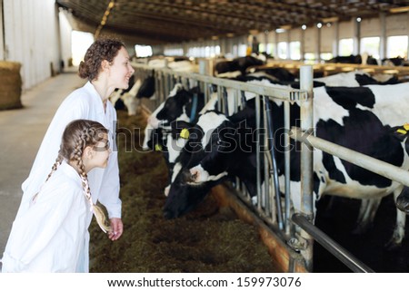 Smiling mother and little daughter look at many cows in stall.