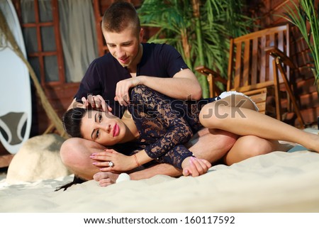 Young man sits on sand and beautiful woman lies on his lap next to beach house.