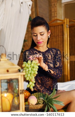 Pretty woman sits on bed and holds bunch of green grapes in bedroom.